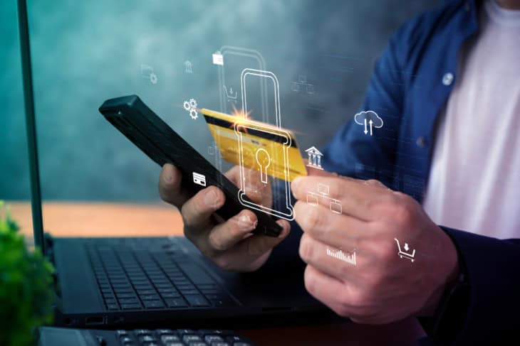close-up of hands using a smartphone and credit card above a laptop with a digital lock overlay, illustrating secure online payment, fintech protection, ecommerce checkout, and mobile banking