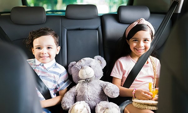 Sister and brother and teddy bear in the back seat of a vehicle
